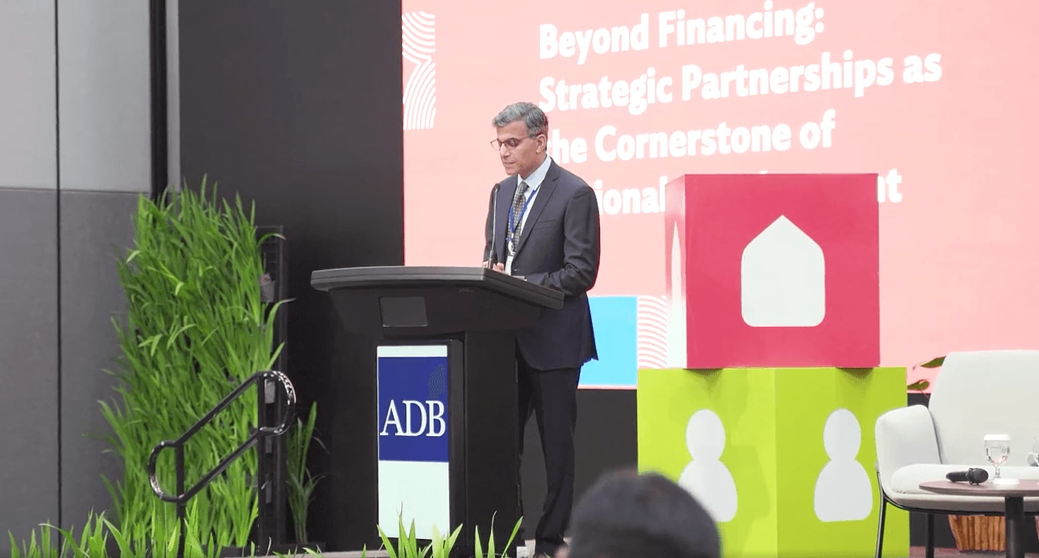A speaker at an event discussing strategic partnerships in development, with a colorful backdrop and plants.