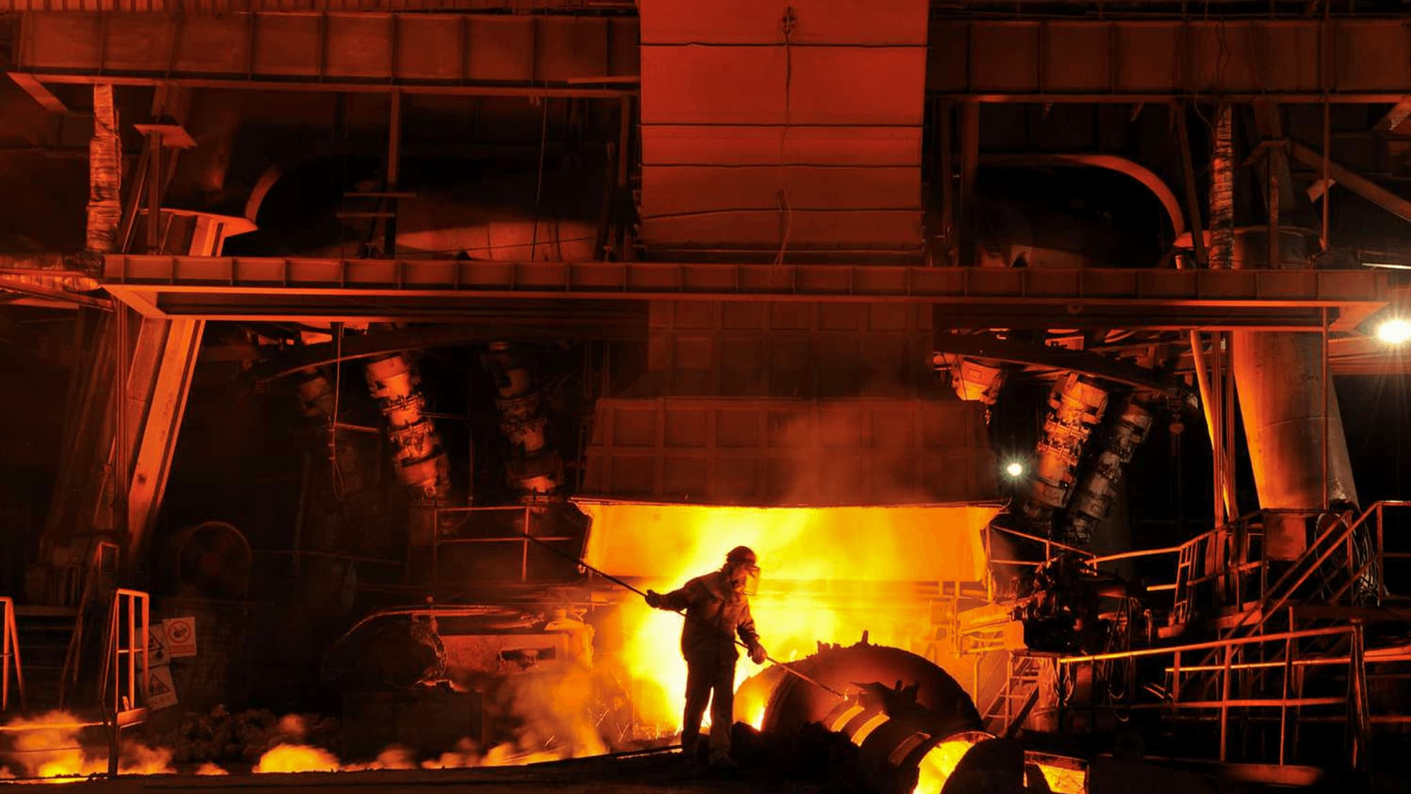 Worker in steel mill casting molten metal, surrounded by glowing furnaces and industrial machinery.