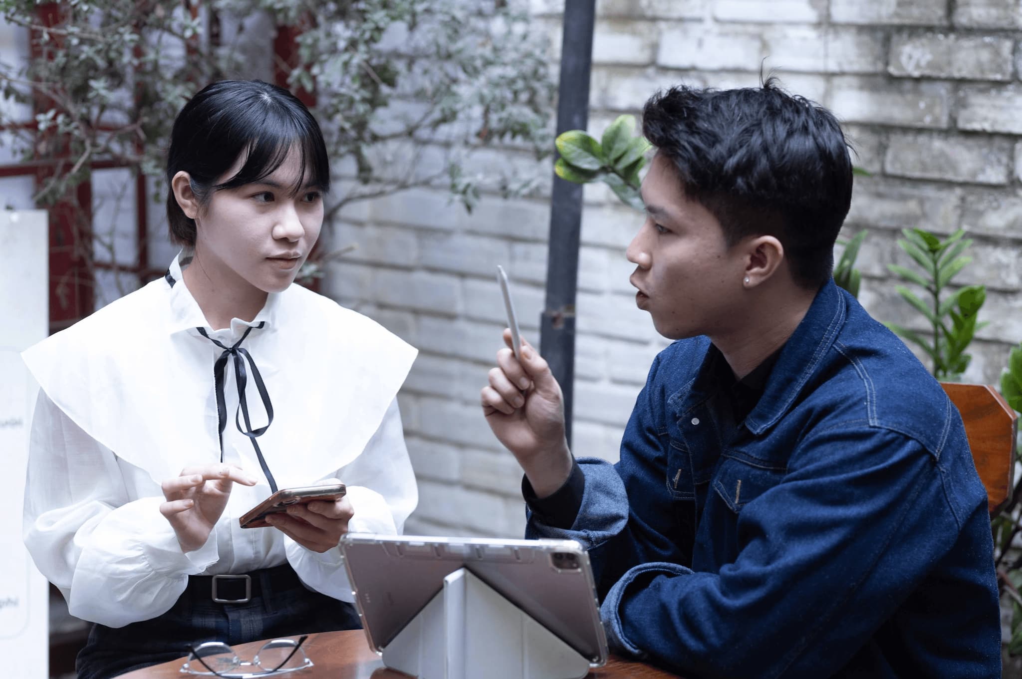 Two young professionals engaged in a discussion at a café, one using a tablet and the other holding a smartphone.