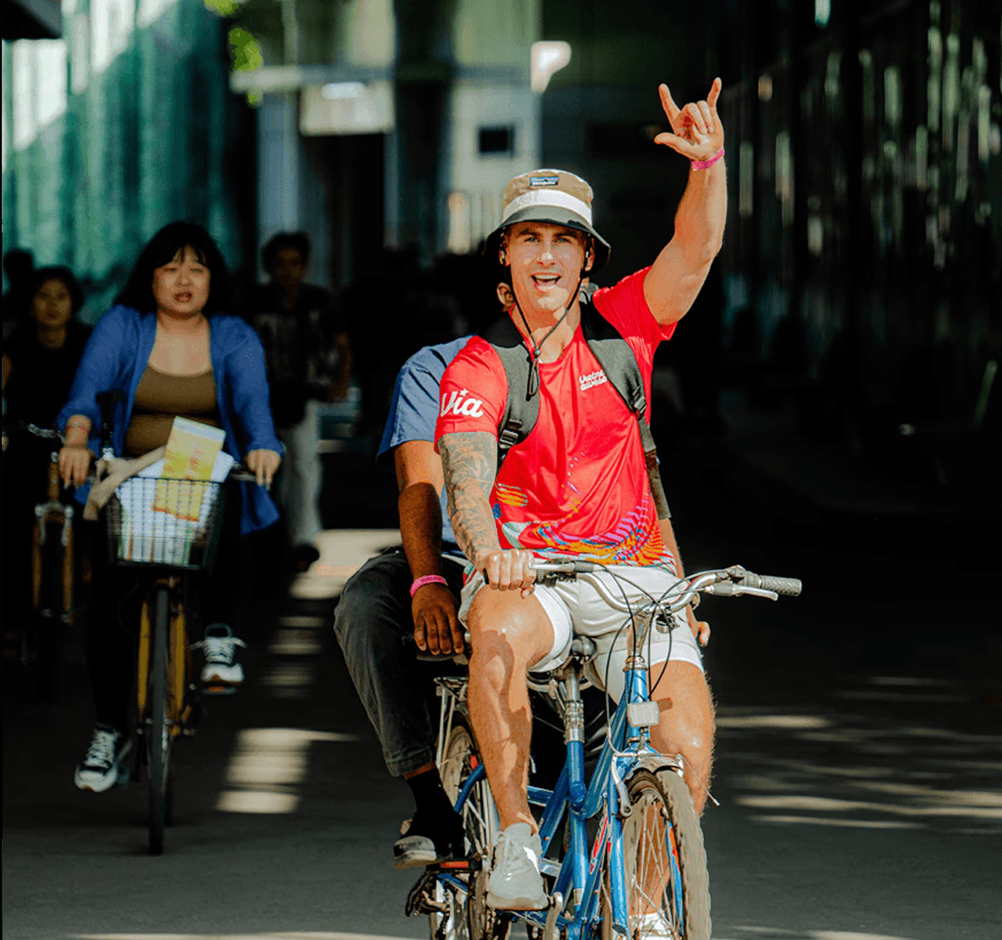 A cyclist wearing a red shirt and hat raises a hand while riding through a bustling urban area with other bikers.