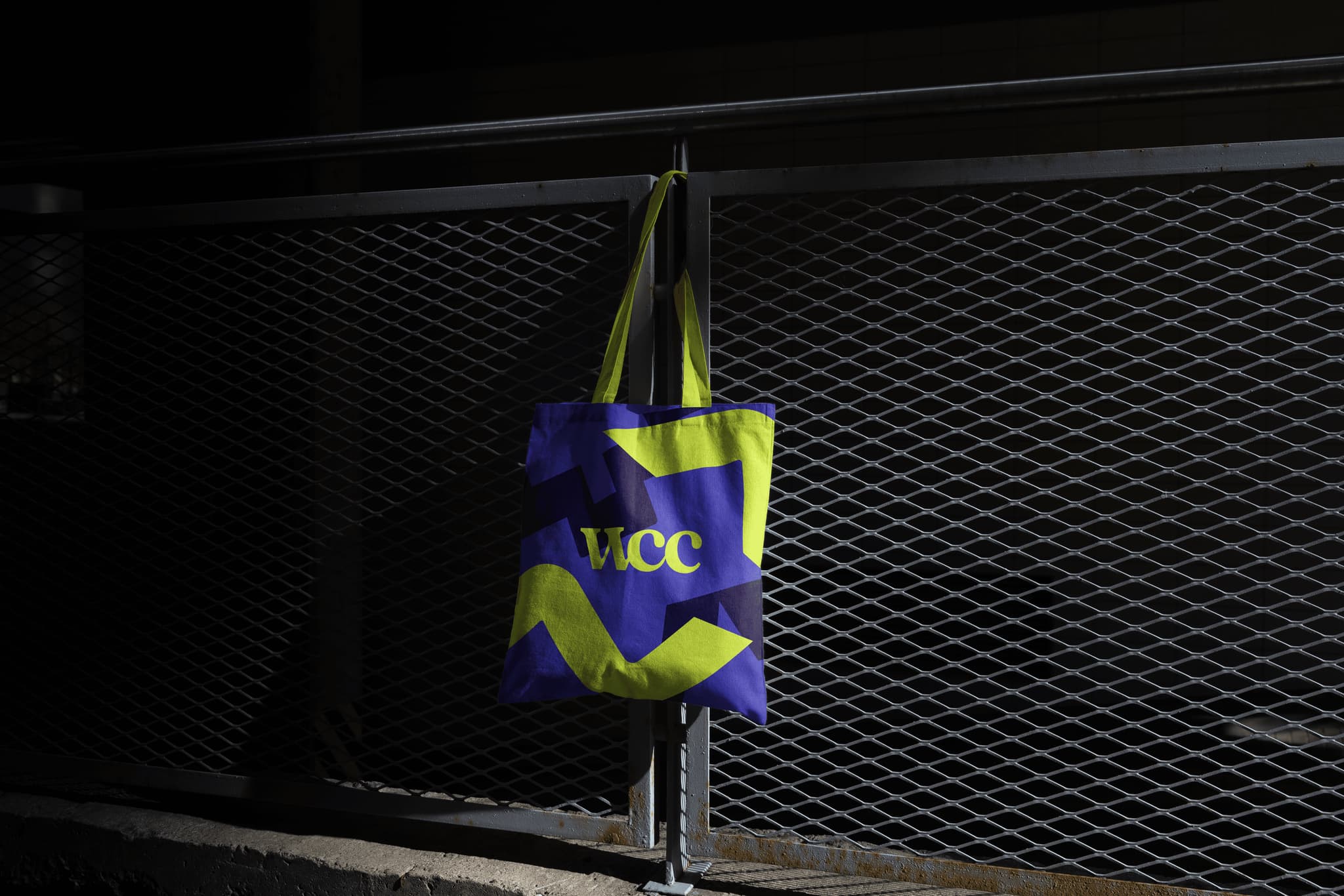VCC tote bag in purple and yellow hanging on a metal fence at night, showcasing a vibrant design.
